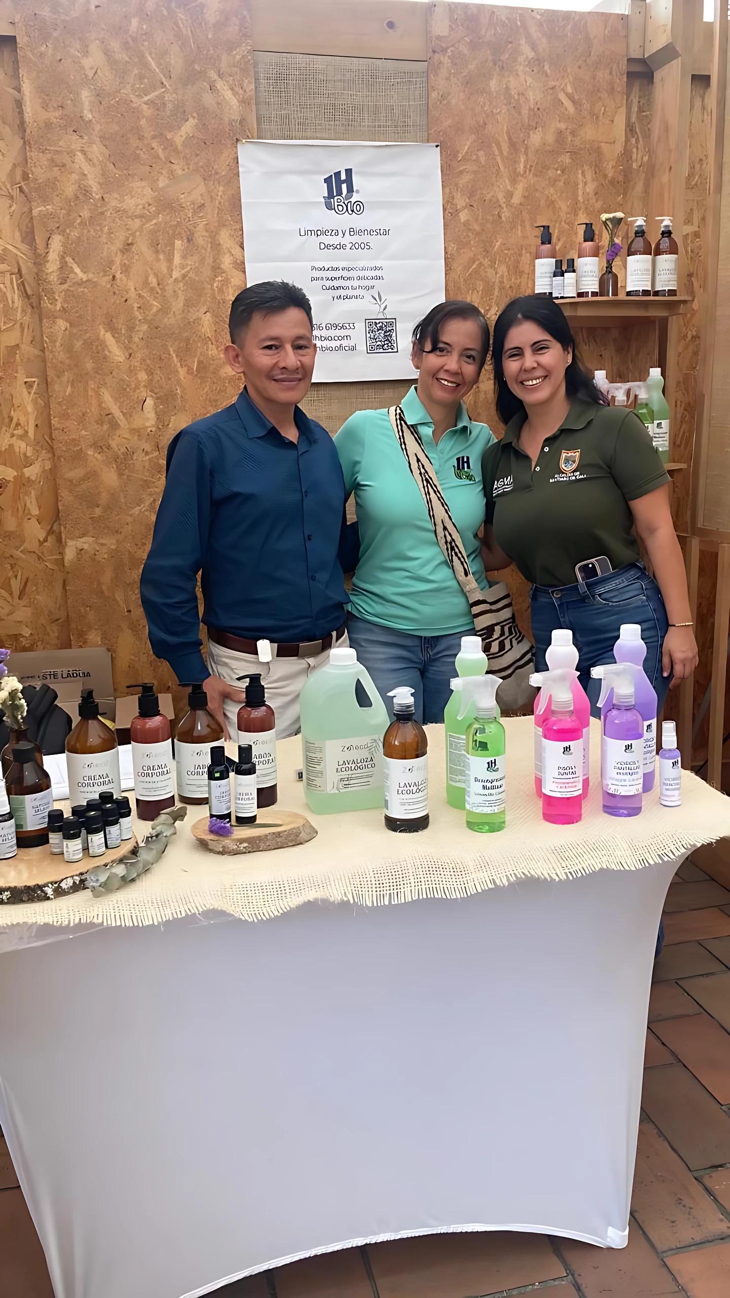 Tres personas posan frente a una mesa con jabones corporales naturales Zoieco en botellas de vidrio transparente con etiquetas blancas y tapas de colores, sobre mantel blanco con borla.