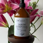 Botella de jabón corporal natural Zoieco con extracto de coco orgánico, color ámbar, sobre pedestal oscuro, rodeada de lirios rosados.