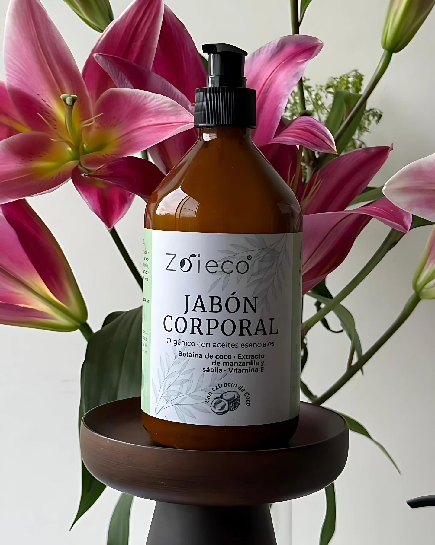 Botella de jabón corporal natural Zoieco con extracto de coco orgánico, color ámbar, sobre pedestal oscuro, rodeada de lirios rosados.