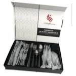Juego de cubiertos acero inoxidable negro mate, caja abierta mostrando 24 piezas individuales en compartimentos, con tenedores, cuchillos y cucharas envueltas en plástico transparente.