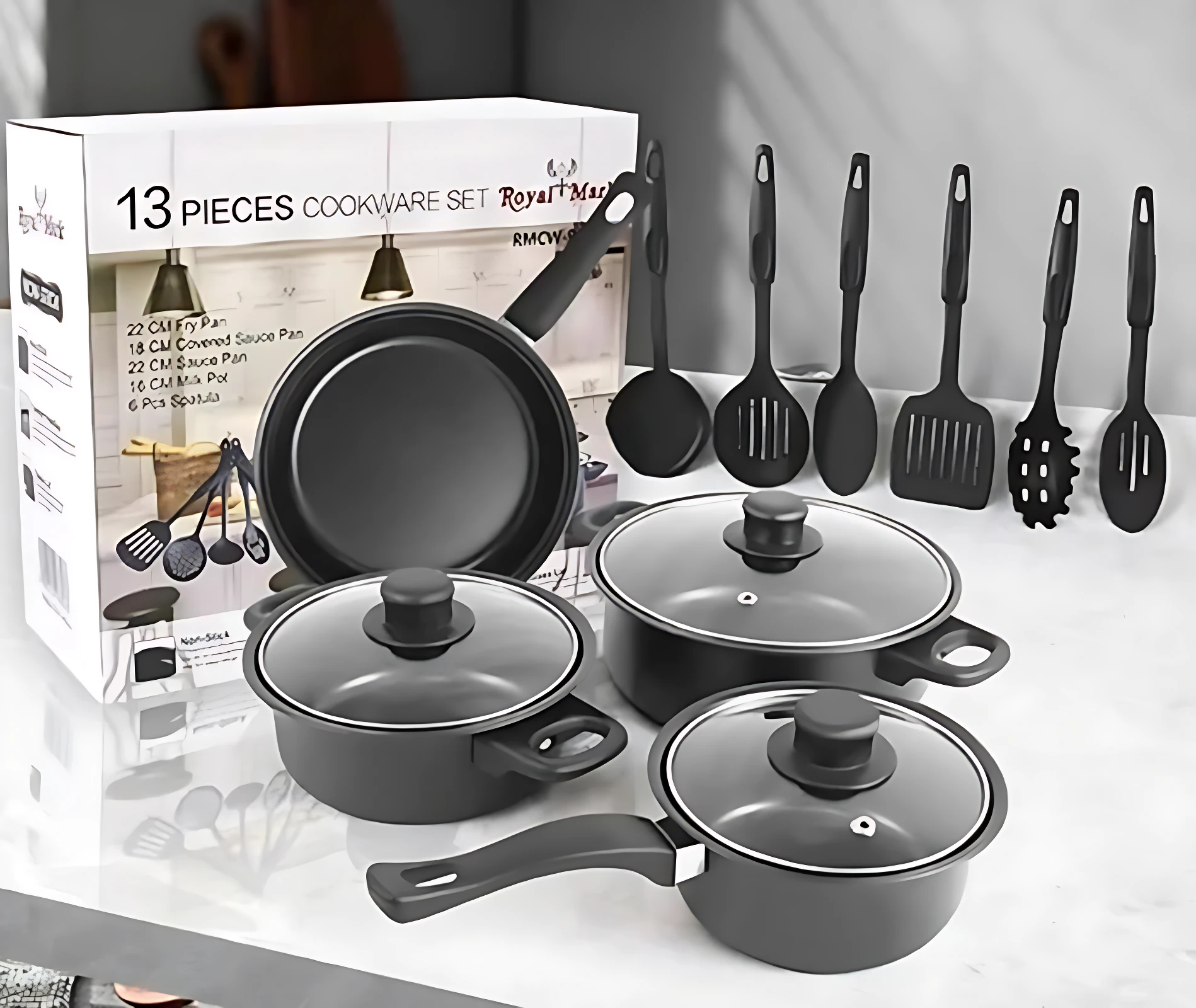 Juego de ollas negras de 13 piezas con tapas de vidrio y utensilios de cocina, sobre superficie blanca junto a su caja.