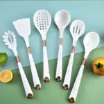 Set de siete utensilios de cocina en silicona blanca con detalles dorados, incluyendo espátula, colador, cuchara y tenedor, sobre fondo verde y azul claro.