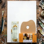 Vista cenital de kit de pintura al óleo sobre mesa de madera con manchas de pintura. Incluye bastidor blanco de 60x40 cm, pinceles nuevos en empaque, paleta de madera, tubos de pintura y trapos usados.