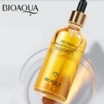 BIOAQUA, sérum de 24K Gold Hyaluronic Essence en frasco de vidrio transparente con líquido dorado y gotas visibles, sobre superficie curva reflectante.