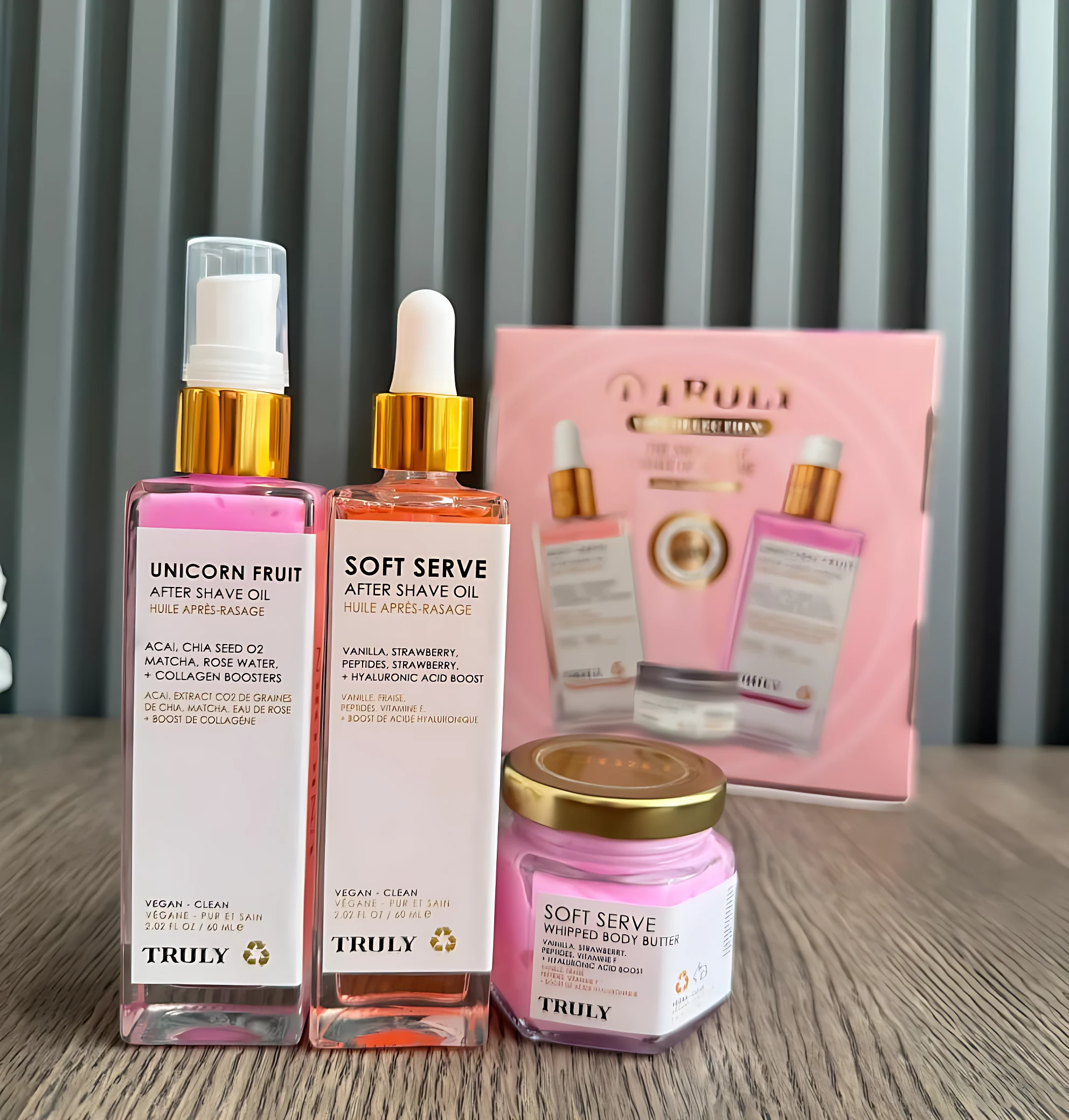 Kit de cuidado corporal Truly con dos aceites después de afeitado, un bálsamo corporal y su caja rosa, sobre superficie de madera.