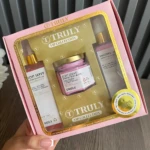 Kit de cuidado corporal Truly en caja rosa con tres productos: aceite de cuerpo, crema hidratante y loción corporal, todos con etiquetas doradas y tapas metálicas.