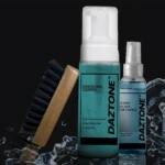 Kit de limpieza Daztone para calzado: cepillo de cerdas negras con mango madera, spray azul translúcido y botella de espuma azul con tapa blanca, sobre fondo negro con salpicaduras de agua.