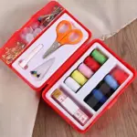Kit de costura compacto en caja roja abierta, con tijeras naranjas, hilos multicolor, agujas, botones, cinta métrica y alfileres organizados en compartimentos blancos.