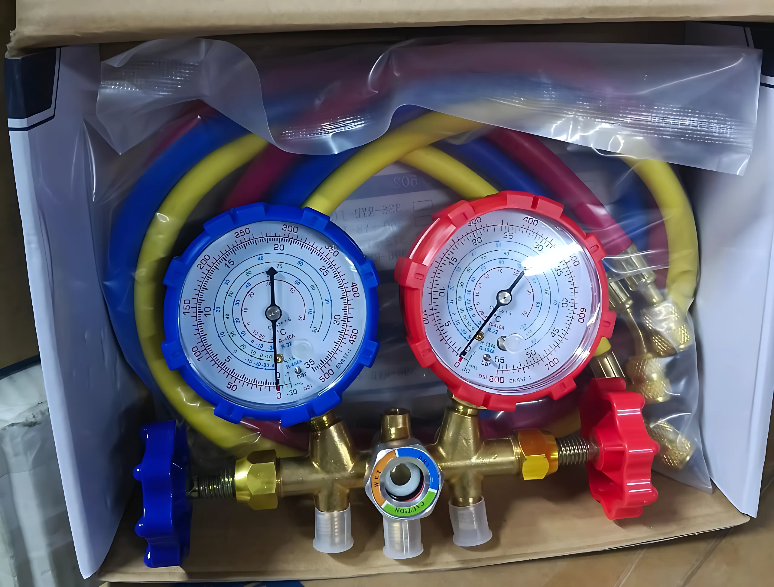 Kit de manómetros de refrigeración con dos indicadores, uno azul y otro rojo, conectados a mangueras amarillas y rojas, junto a válvulas de control en bronce y llaves de paso en colores azul y rojo, todo contenido en caja con protección plástica.