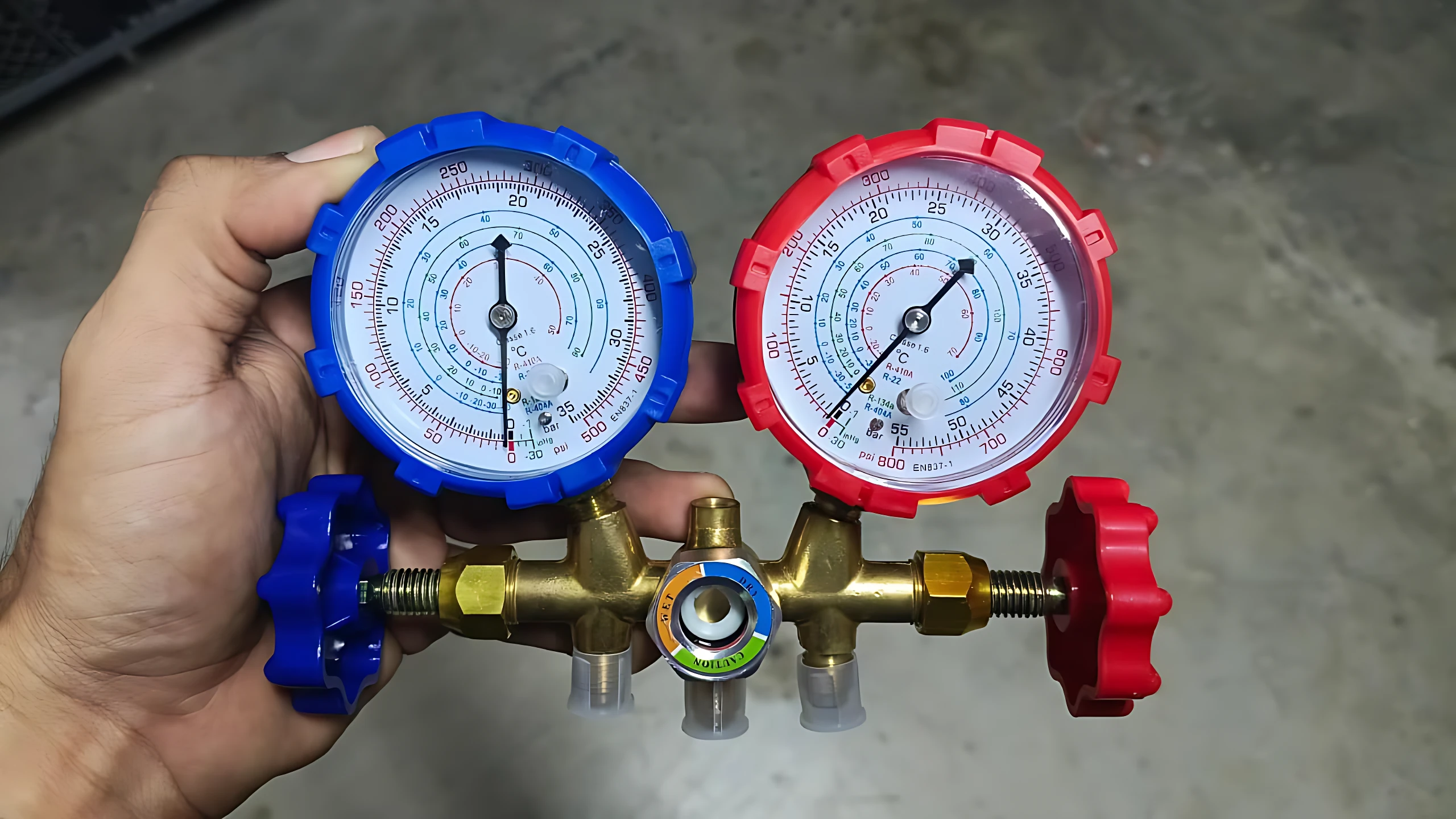 Kit de manómetros para refrigeración con dos medidores de presión en azul y rojo, conectados a una válvula central dorada con llaves rojas y azules, diseñado para diagnóstico de fugas y carga de refrigerante.