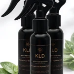 Tres botellas negras de spray termoactivo Kld con etiquetas doradas, dispuestas en diagonal sobre fondo blanco, acompañadas de hojas verdes frescas a los lados.