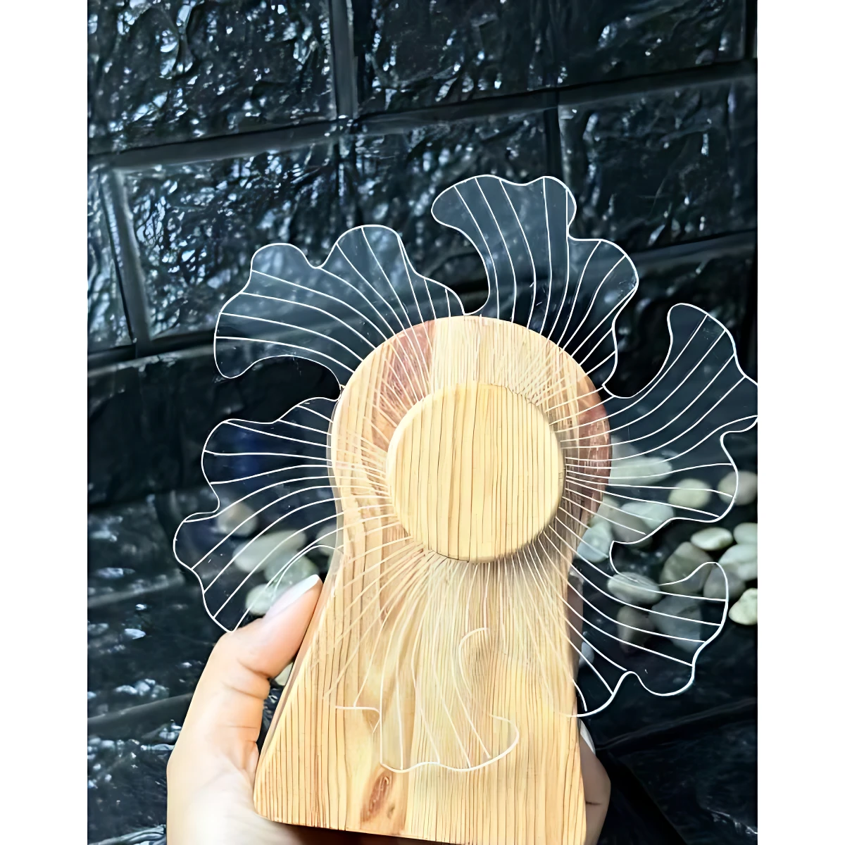 Lámpara 3D giratoria de madera con base circular y aletas acrílicas transparentes, sostenida en mano frente a fondo de baldosas oscuras.