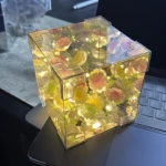Cubo de luz LED con efecto espejo y girasoles internos, colocado sobre teclado de laptop.