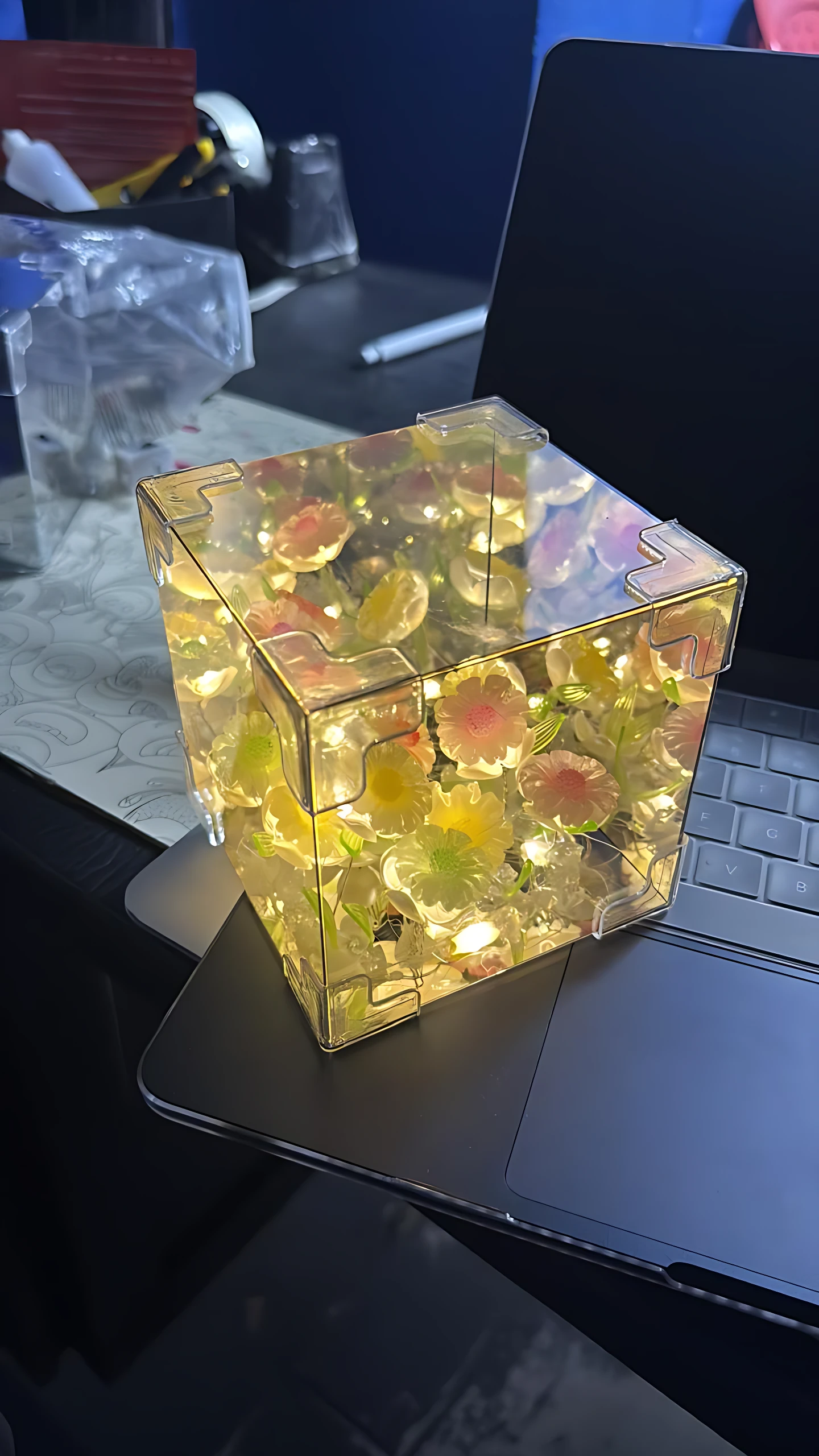 Cubo de luz LED con efecto espejo y girasoles internos, colocado sobre teclado de laptop.