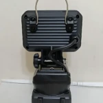 Vista trasera de la lámpara de trabajo inalámbrica 1 Bat, color negro mate, con asa metálica superior, panel de ventilación trasero y cable de carga conectado al soporte ajustable.