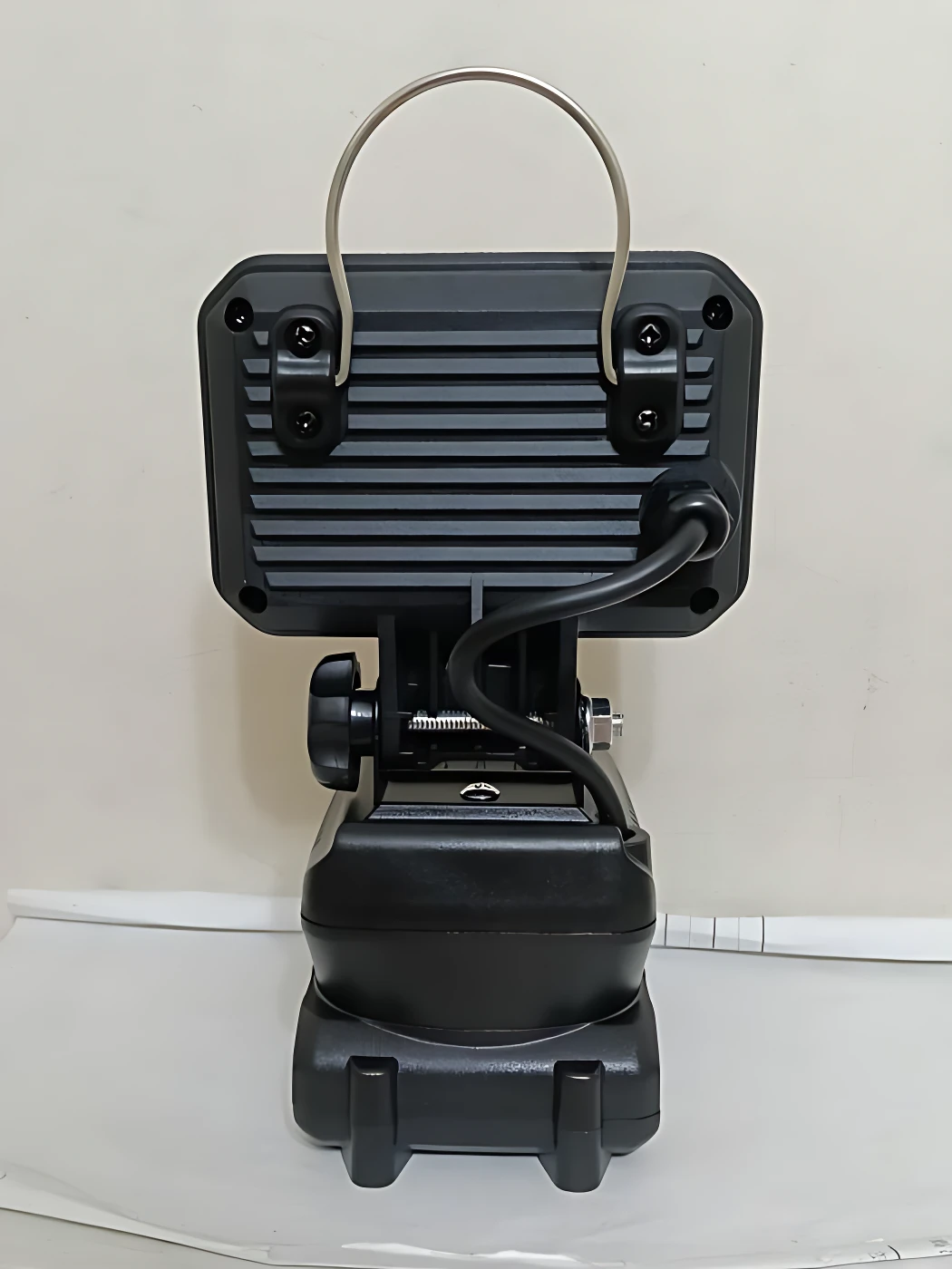 Vista trasera de la lámpara de trabajo inalámbrica 1 Bat, color negro mate, con asa metálica superior, panel de ventilación trasero y cable de carga conectado al soporte ajustable.