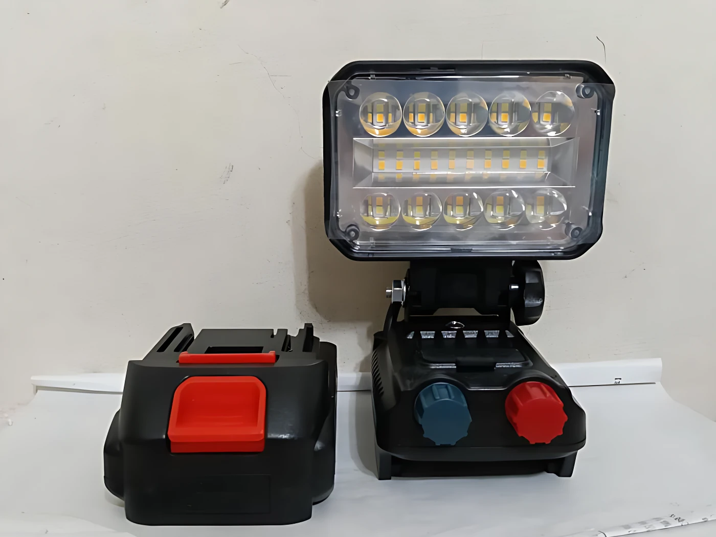 Lámpara de trabajo inalámbrica con batería incluida, panel LED frontal y controles rotativos en base negra.