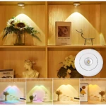 Lámpara LED circular blanca con luz cálida, instalada en estantería de madera; incluye variante RGB y vista frontal del módulo.