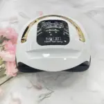 Lámpara de uñas LED 168W en color blanco con detalles dorados, vista frontal sobre superficie de mármol con flores rosadas al lado.