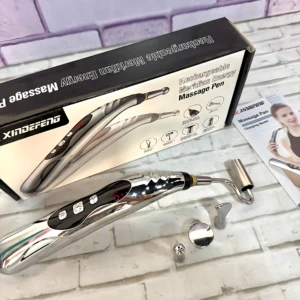 Lápiz masajeador eléctrico recargable con cabezales intercambiables, cuerpo metálico brillante y caja de presentación sobre fondo de ladrillo blanco.