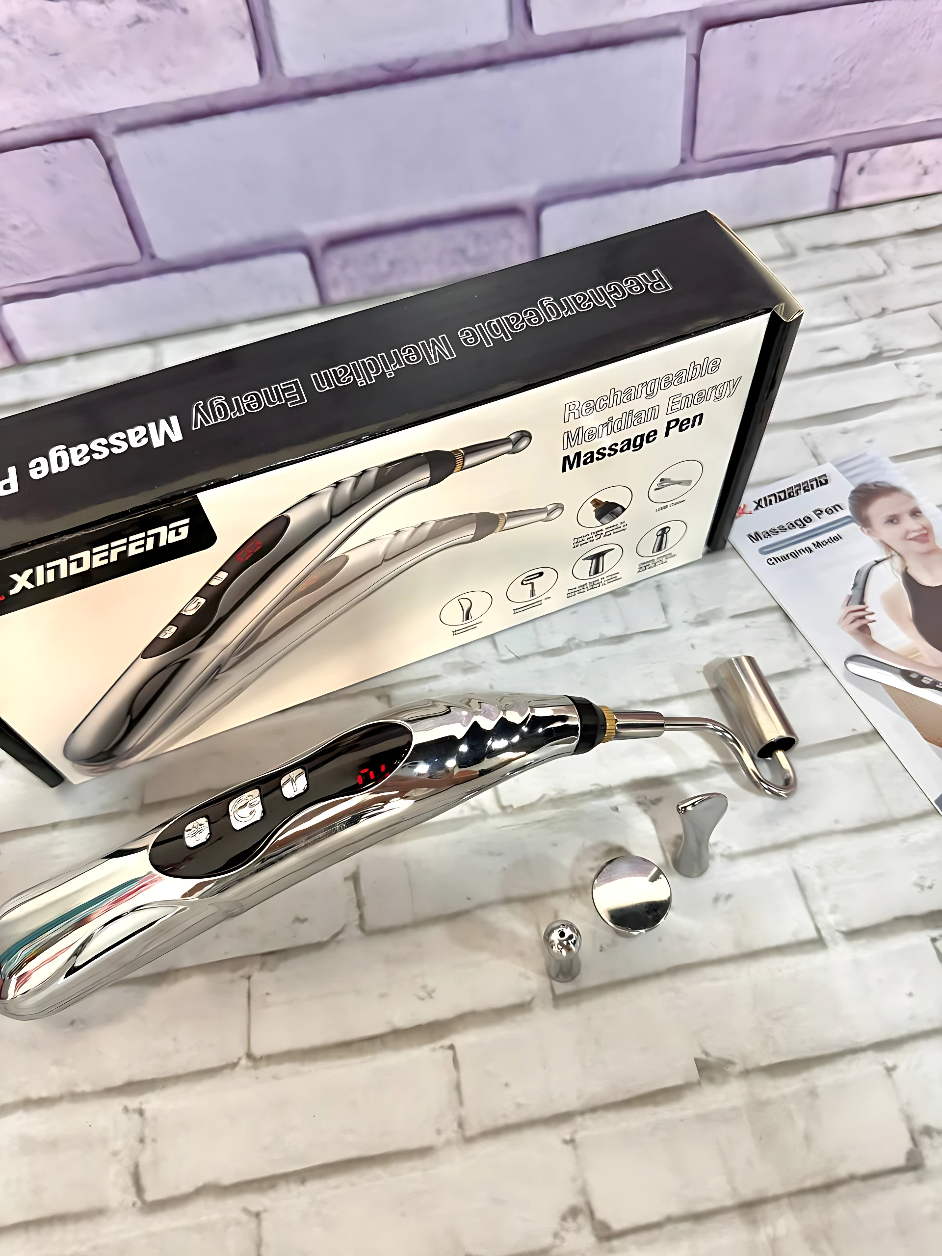 Lápiz masajeador eléctrico recargable con cabezales intercambiables, cuerpo metálico brillante y caja de presentación sobre fondo de ladrillo blanco.