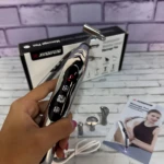 Lápiz masajeador eléctrico recargable en mano, con cabezales intercambiables y caja de presentación al fondo sobre superficie blanca.