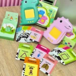 Lector de inglés interactivo para niños en colores verde y rosa, con tarjetas de vocabulario en español e inglés sobre mesa de madera.