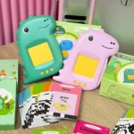 Dos lectores interactivos para niños en forma de dinosaurio, uno verde y otro rosa, con pantalla amarilla. Incluyen tarjetas de vocabulario en inglés y español, como 'grey' y 'triangle', sobre una mesa de madera.