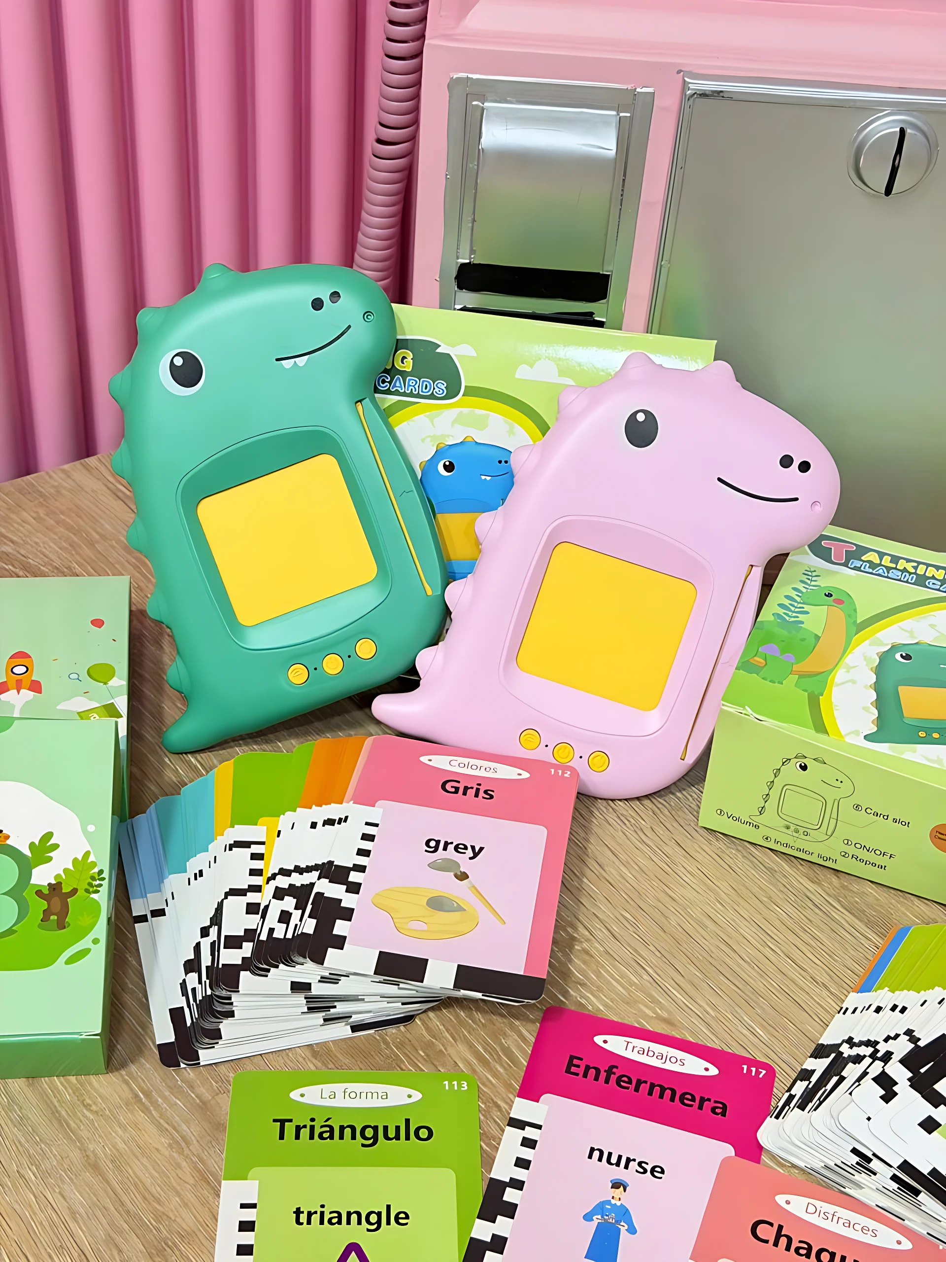 Dos lectores interactivos para niños en forma de dinosaurio, uno verde y otro rosa, con pantalla amarilla. Incluyen tarjetas de vocabulario en inglés y español, como 'grey' y 'triangle', sobre una mesa de madera.