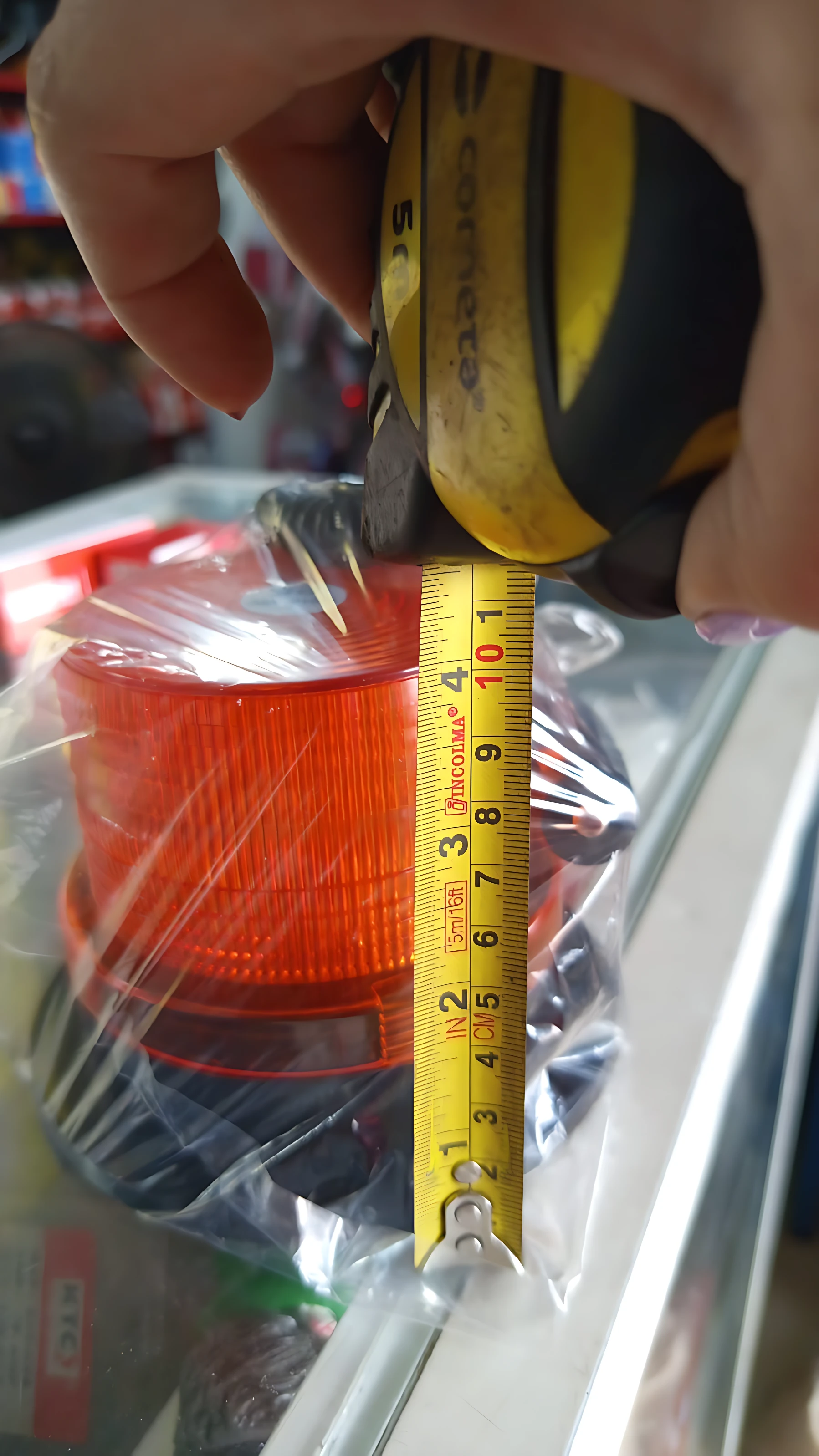 Luz de advertencia naranja con carcasa plástica, envuelta en plástico transparente, siendo medida con cinta métrica amarilla que indica aproximadamente 10 centímetros de altura.