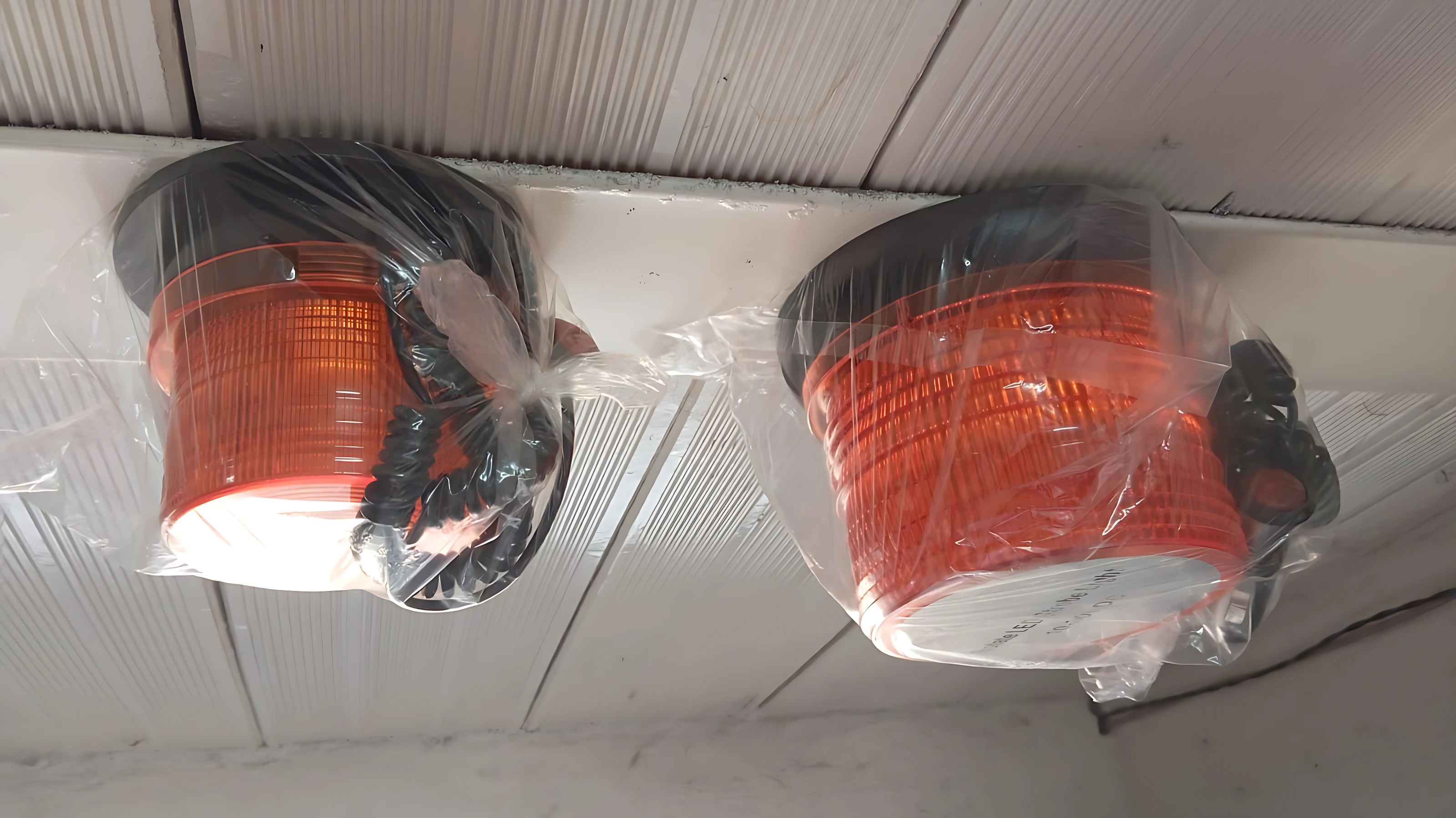 Dos luces de advertencia naranjas con base negra, envueltas en plástico transparente, montadas sobre techo de paneles blancos.