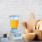 Licuadora multifuncional de 600W con jarra de vidrio azul claro, base blanca y panel de control con botones. Incluye accesorios de madera: cuchara, tenedor, bowl y canasta. Fondo de ladrillo blanco.