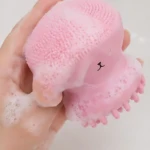 Limpiador facial de silicona en forma de pulpo, color rosa, con textura de cerdas suaves y detalles de masaje; se muestra en uso con espuma.
