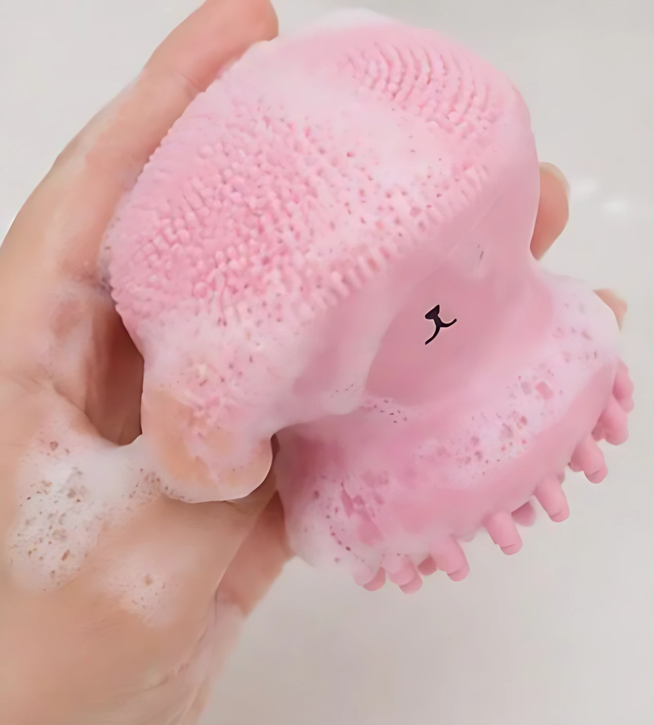 Limpiador facial de silicona en forma de pulpo, color rosa, con textura de cerdas suaves y detalles de masaje; se muestra en uso con espuma.