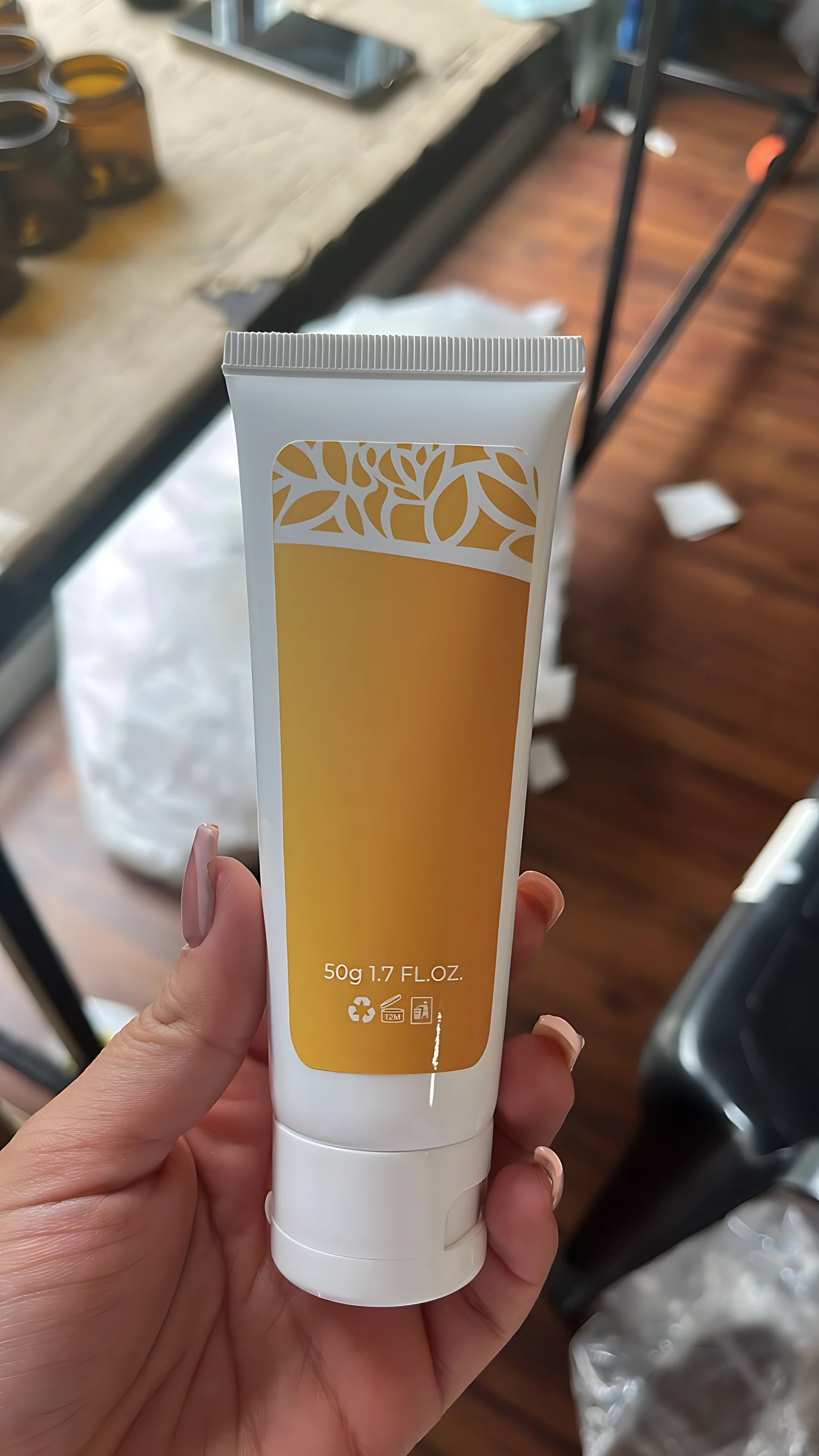 Tubo de exfoliante corporal Lumina en color amarillo con diseño floral, contenido de 50g, sostenido en mano sobre fondo de madera.