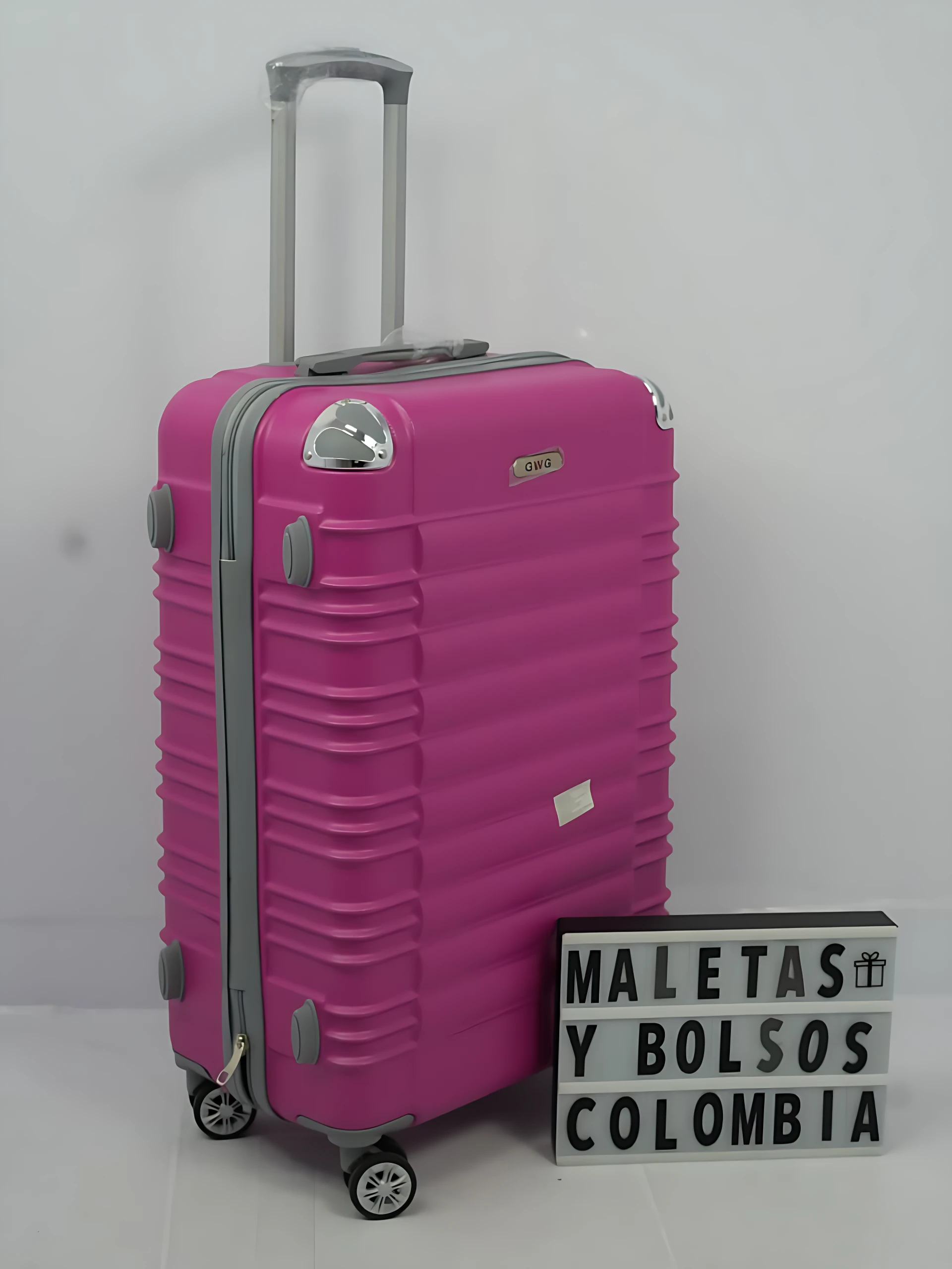 Maleta rígida rosa con ruedas giratorias y asa telescópica, junto a letrero que dice MALETAS Y BOLSOS COLOMBIA.