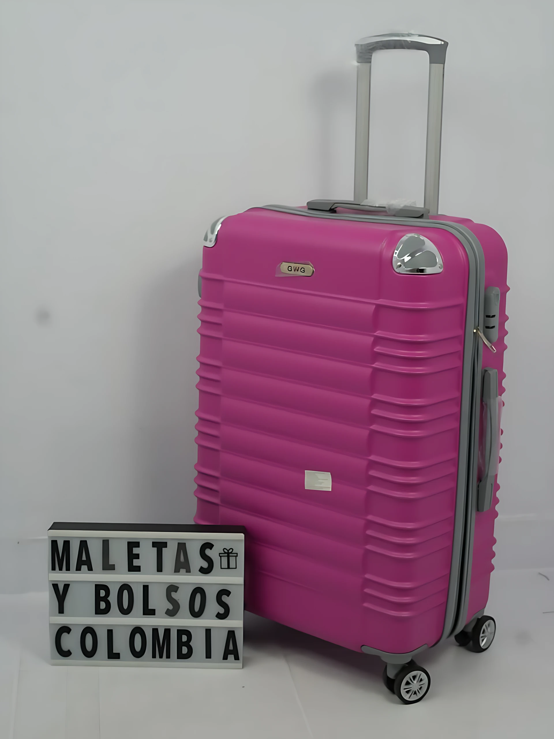 Maleta rígida rosa con ruedas giratorias y asa telescópica, junto a un letrero que dice "MALETAS Y BOLSOS COLOMBIA".