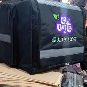Maleta negra de La Uvita con logo visible, logotipo en morado y verde, número de contacto impreso, sobre superficie con maletas y mochilas al fondo.