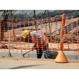Malla naranja de polipropileno para señalización, instalada en obra con postes amarillos, delimitando zona de trabajo con trabajador en casco y chaleco.