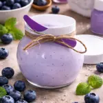 Mantequilla corporal arándanos en frasco de vidrio con tapa blanca y lazo de cuerda, rodeada de frutos frescos y menta.