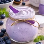 Mantequilla corporal arándanos en frasco de vidrio con tapa, textura cremosa y color lavanda, rodeada de frutos frescos y menta.