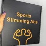Caja negra con texto amarillo que dice Sports Slimming Abs y un dibujo de brazos flexionados, mostrando el empaque del masajeador escultor EMS para tonificar abdomen y brazos.
