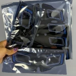Tres masajeadores escultor EMS en bolsas de plástico transparente, mostrando diseño negro con detalles azules y blancos, visibles desde ángulo superior.