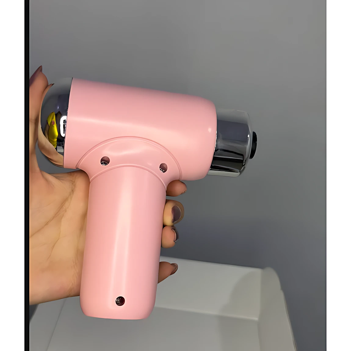 Masajeador percutor portátil en color rosa pastel, con cabezal cromado y mango ergonómico, sostenido en mano para mostrar su tamaño compacto.