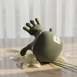 Masajeador eléctrico portátil verde sin cables, con forma de mano y textura suave, sobre superficie clara con flores blancas al fondo.