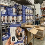Masajeador EMS digital para espalda y cuello, con electrodo azul visible en la caja. Producto en estantería de almacén, con múltiples unidades apiladas.