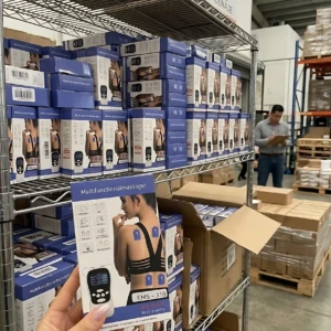 Masajeador EMS digital para espalda y cuello, con electrodo azul visible en la caja. Producto en estantería de almacén, con múltiples unidades apiladas.