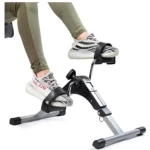 Mini bicicleta spinning compacta en uso, con usuario usando zapatillas blancas y leggings verdes, mostrando pedales ajustables y estructura metálica gris y negra.