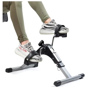 Mini bicicleta spinning compacta en uso, con usuario usando zapatillas blancas y leggings verdes, mostrando pedales ajustables y estructura metálica gris y negra.