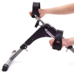 Mini bicicleta spinning compacta en color negro con asideros y correas de seguridad, siendo usada por manos humanas sobre fondo blanco.
